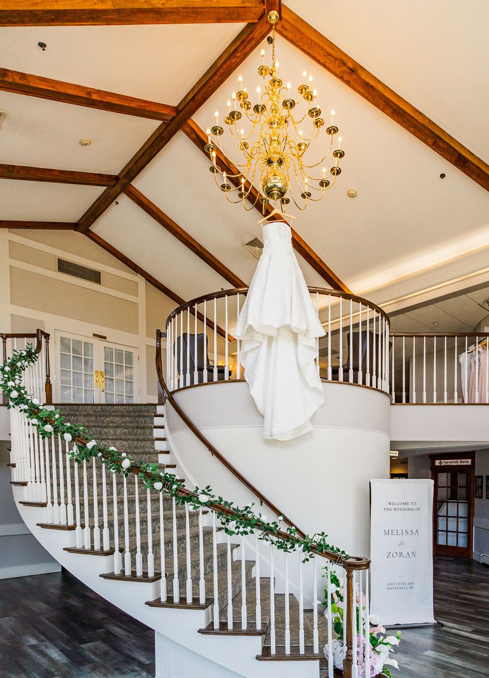 Elegant wedding dress hanging from a grand chandelier above a curved staircase adorned with floral garland.