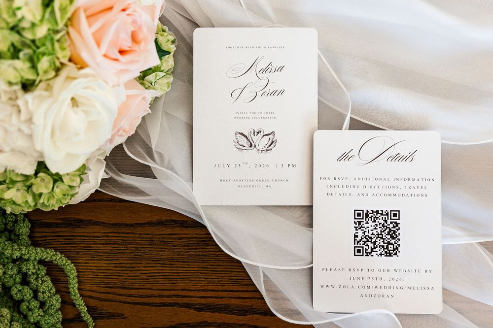 Elegant wedding invitation suite with QR code details card on white veil beside pink and white floral bouquet.