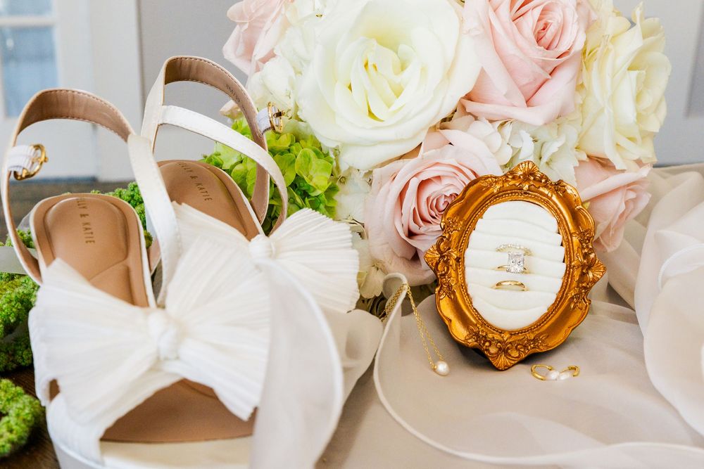 Wedding flat lay featuring white bow heels, gold oval frame with rings, and a blush floral bouquet.