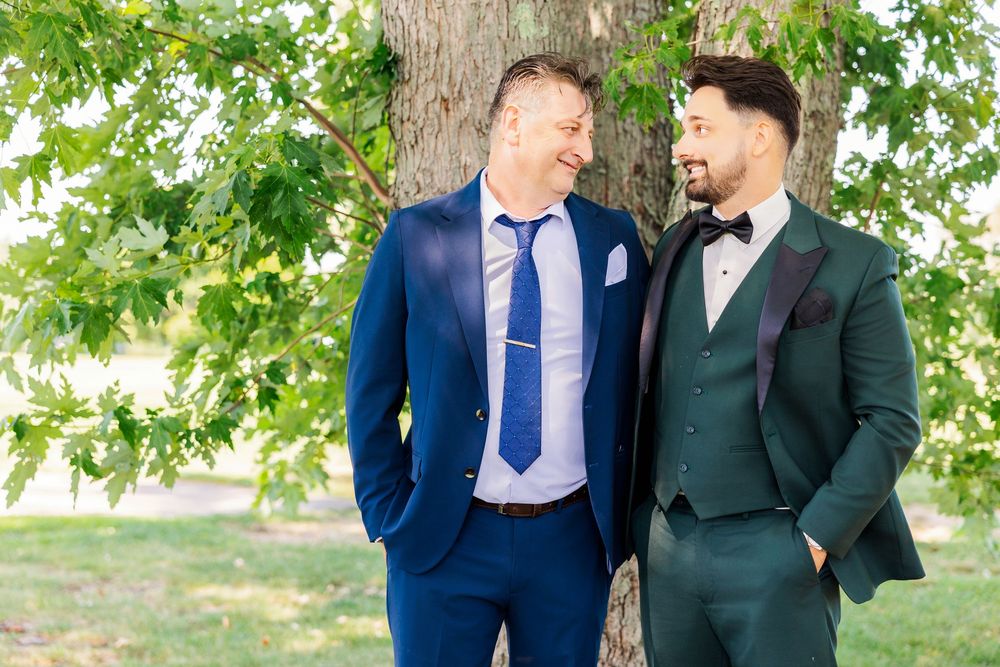 Two grooms in navy and green suits smiling at each other outdoors near a tree on their wedding day.