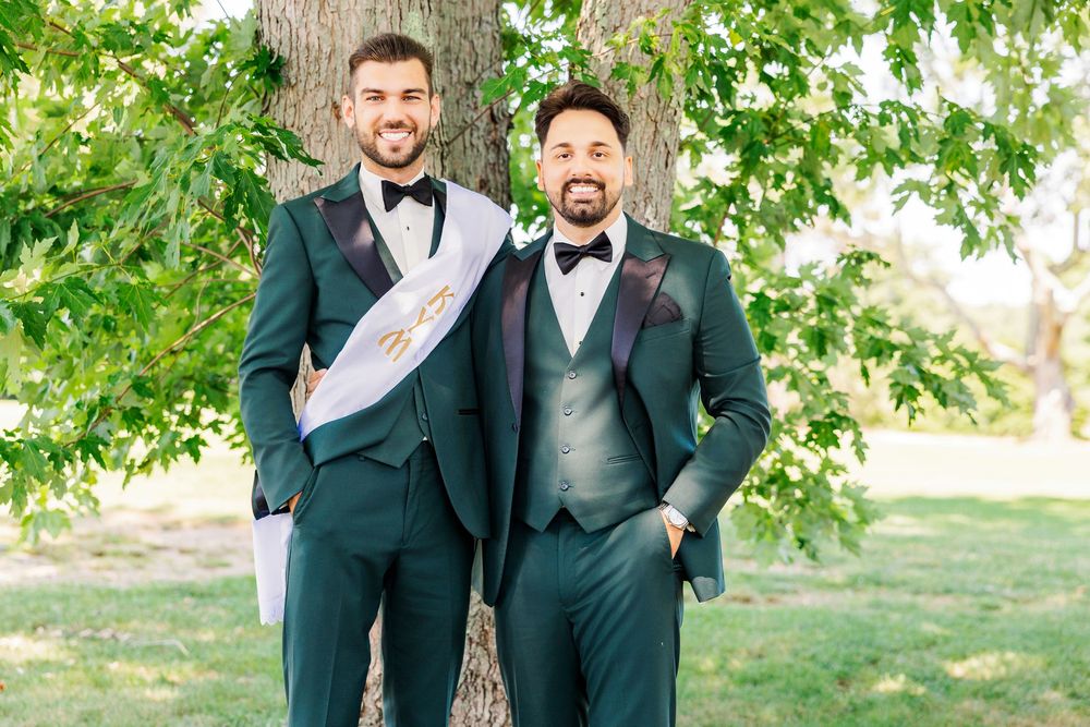 Two grooms in matching dark green tuxedos pose outdoors under a tree on their wedding day.