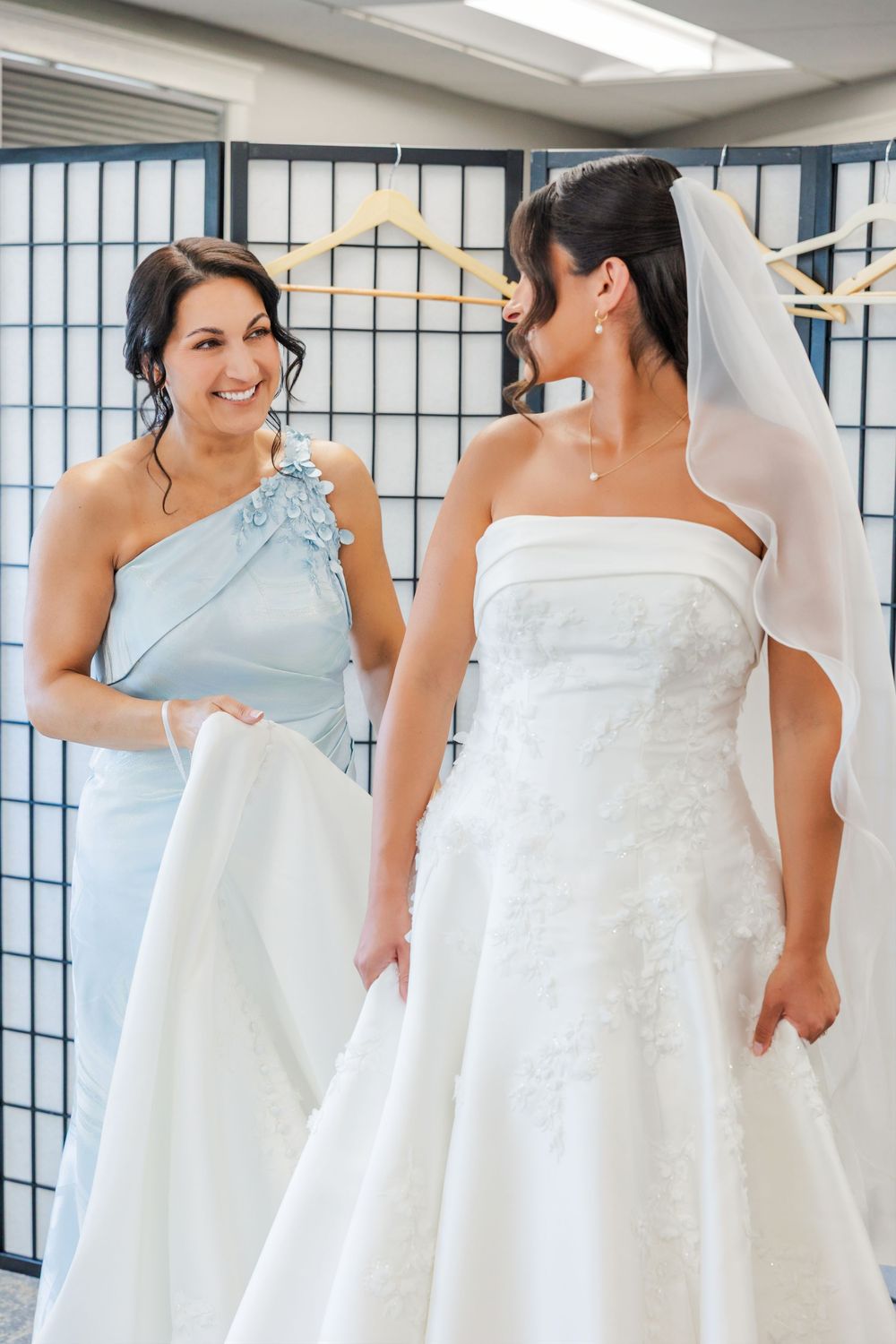 Bridesmaid in light blue dress helps bride in strapless white gown and veil during wedding dress fitting.