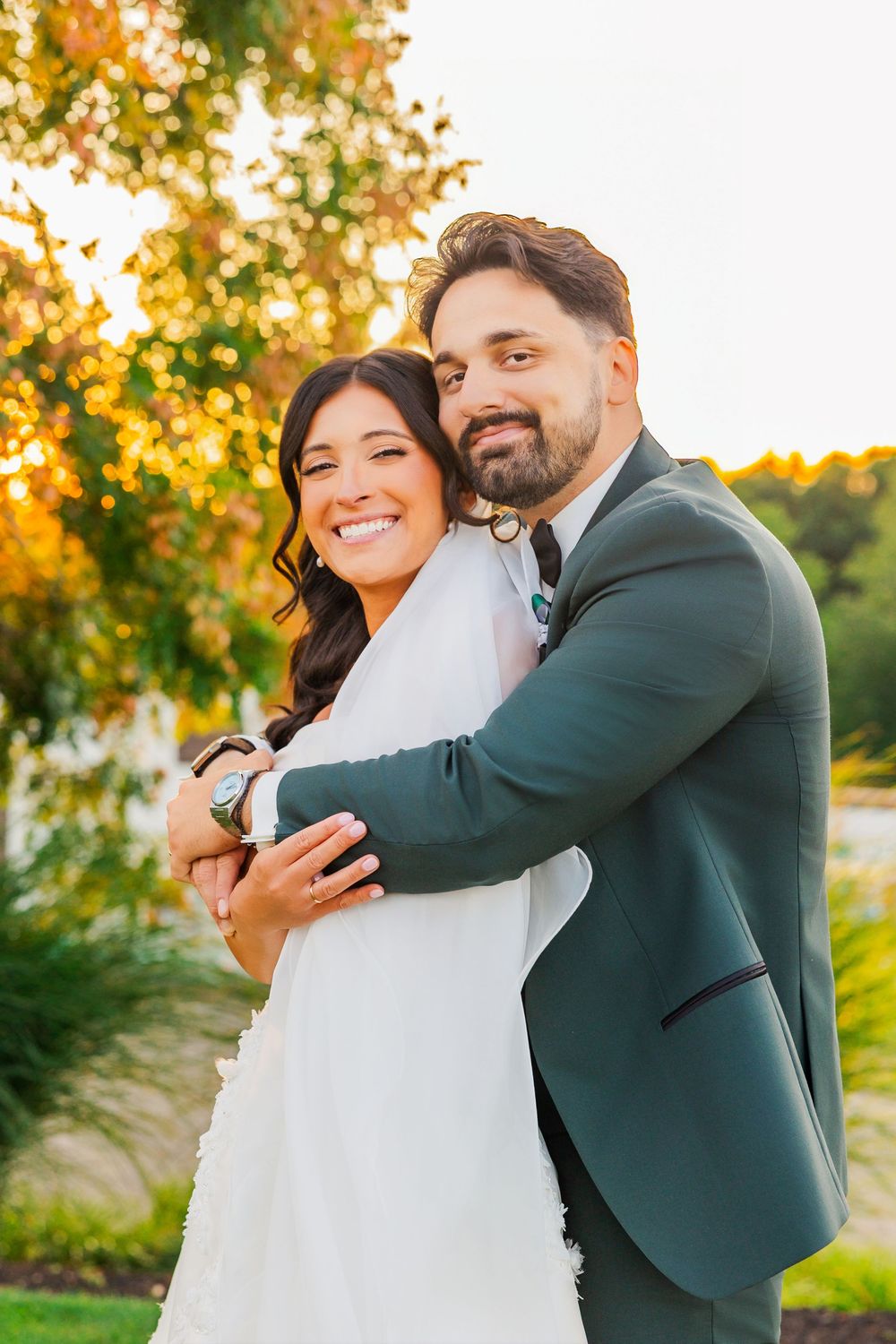 Bride and groom embracing outdoors at golden hour, smiling joyfully in wedding attire with lush greenery behind.