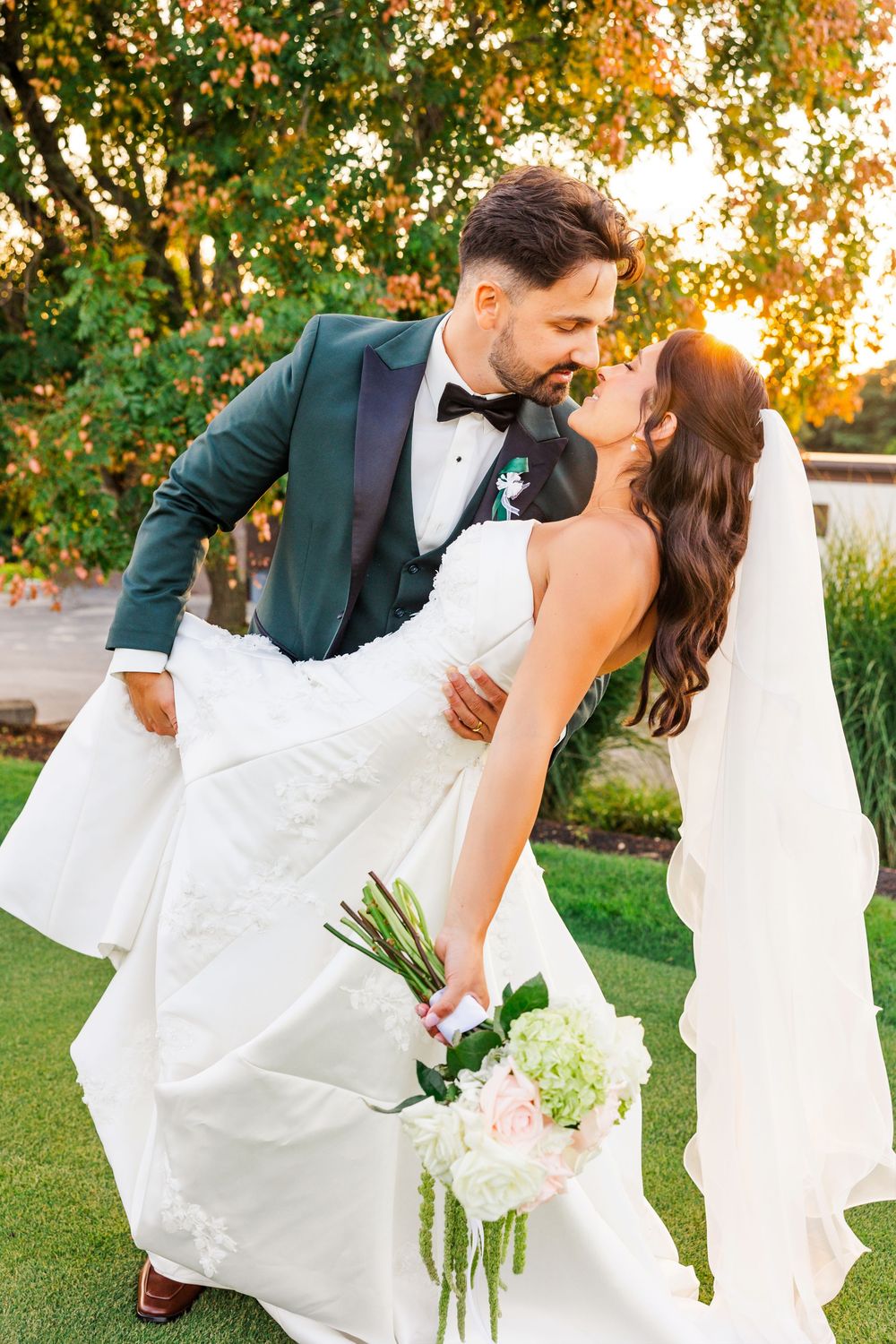 Bride and groom share a romantic dip kiss outdoors at golden hour, bride holding a pink and white bouquet.