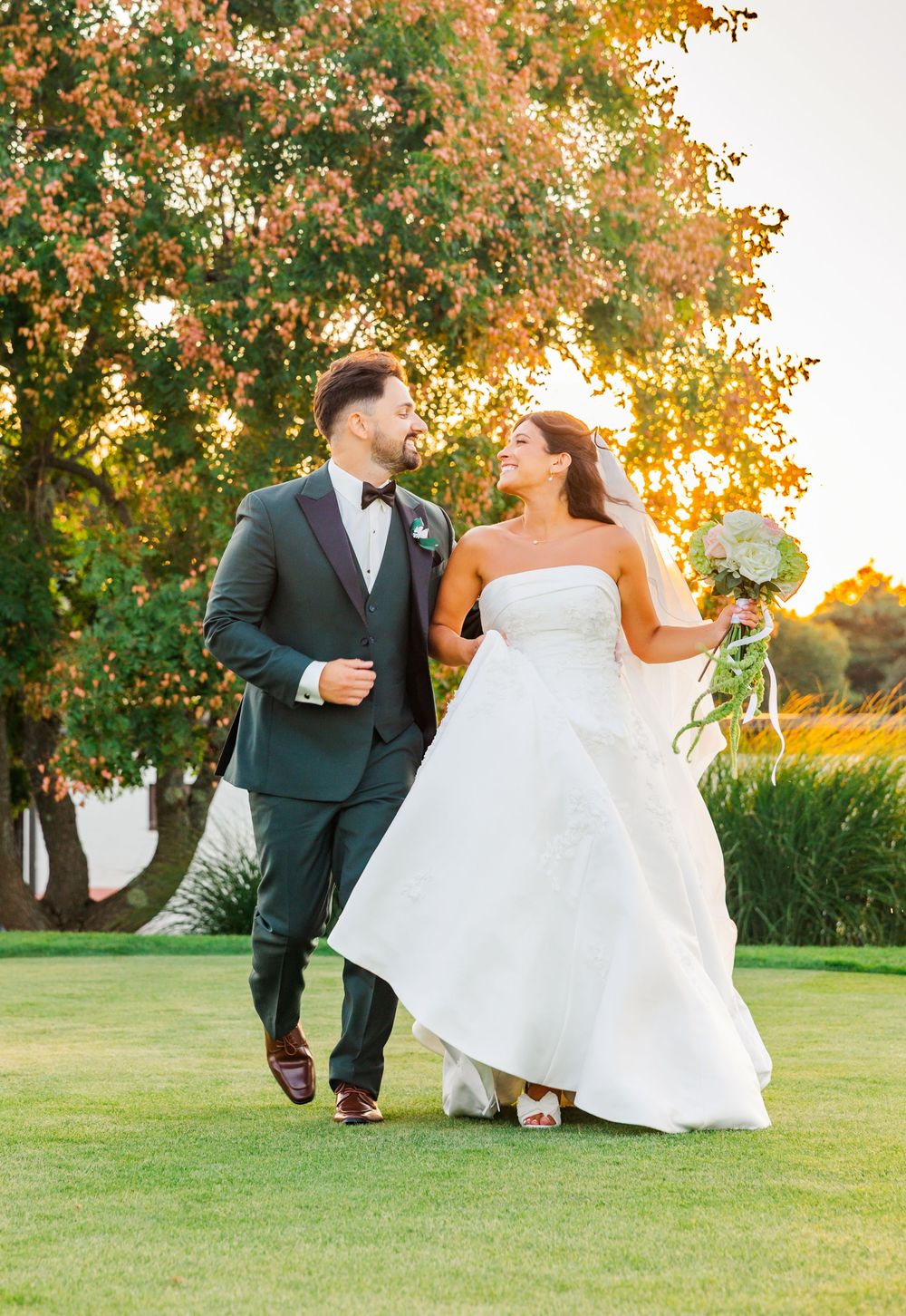 Bride and groom walking on green lawn at golden hour, she holds bouquet, he wears dark green suit with bow tie.