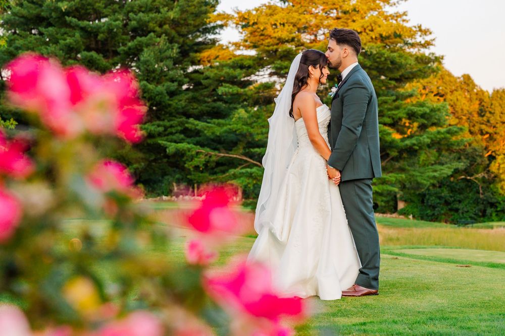 Bride and groom share a romantic moment on lush green lawn with vibrant pink roses and tall trees in background.