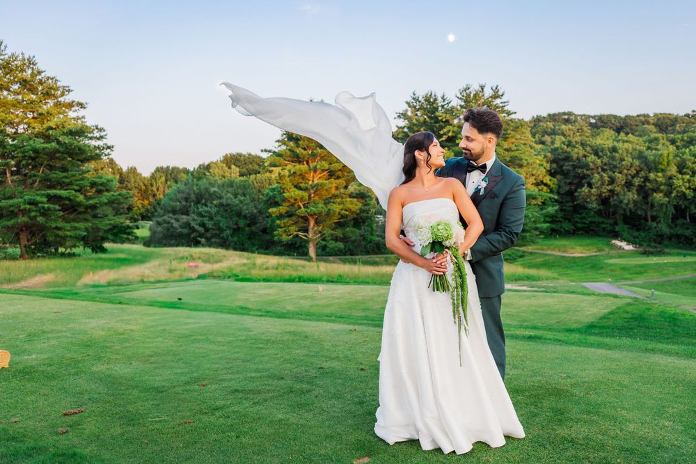 Bride and groom share a romantic moment on a lush golf course, her veil flowing in the breeze.