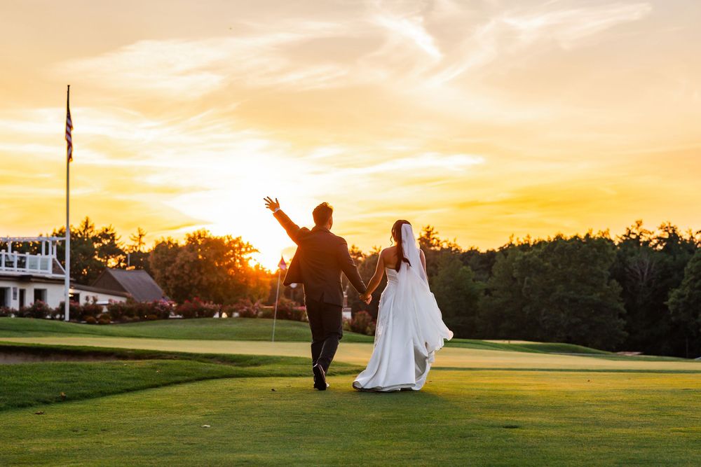 Bride and groom walk hand-in-hand on a golf course at sunset, groom raising his hand in celebration.