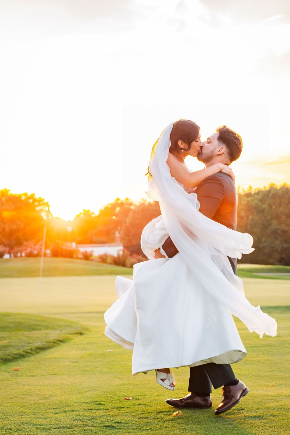 Groom lifts bride in white gown for a romantic kiss at sunset on a lush green golf course.