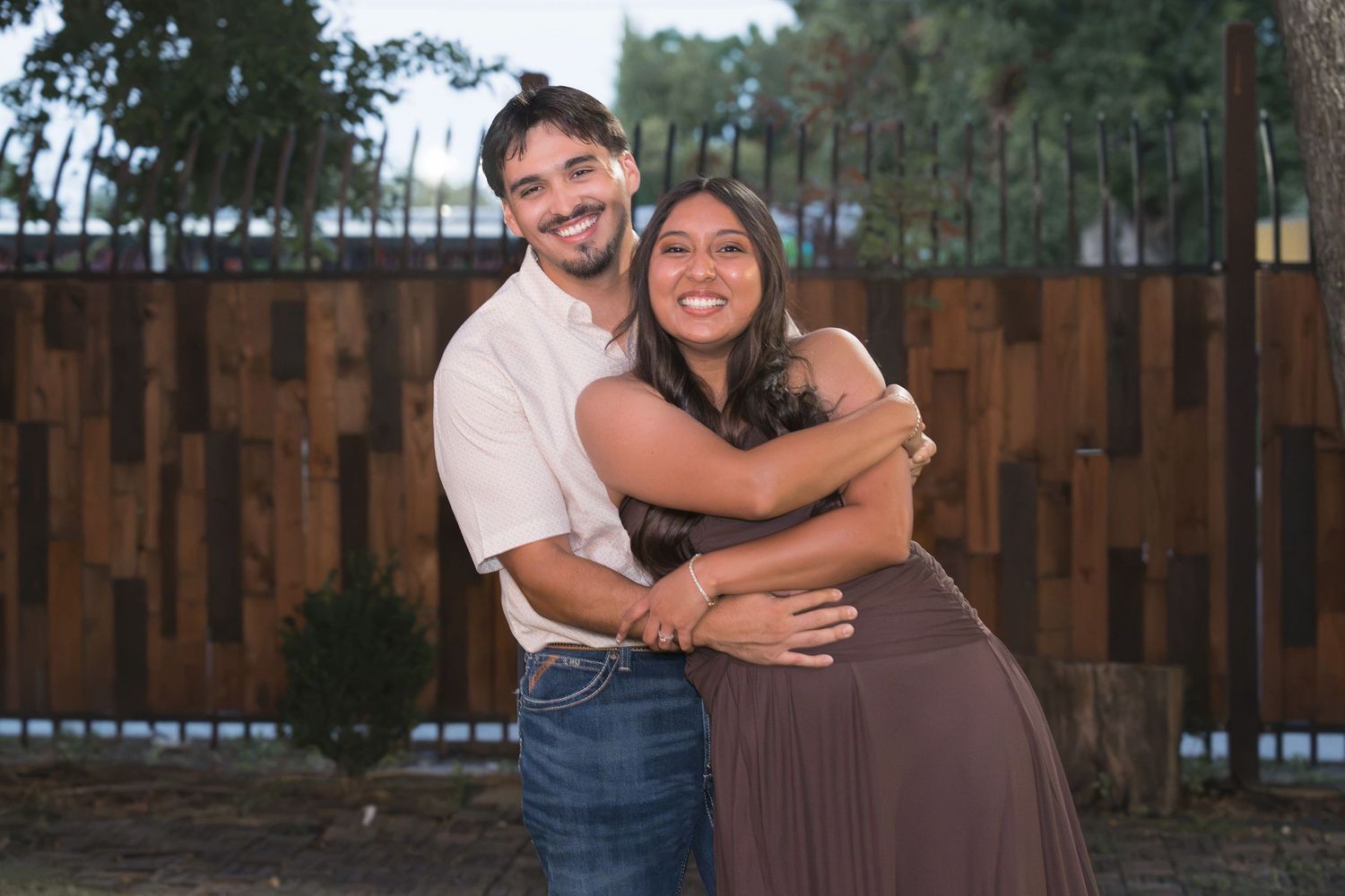 Happy couple embracing outdoors near a wooden fence, smiling warmly in casual attire during golden hour.