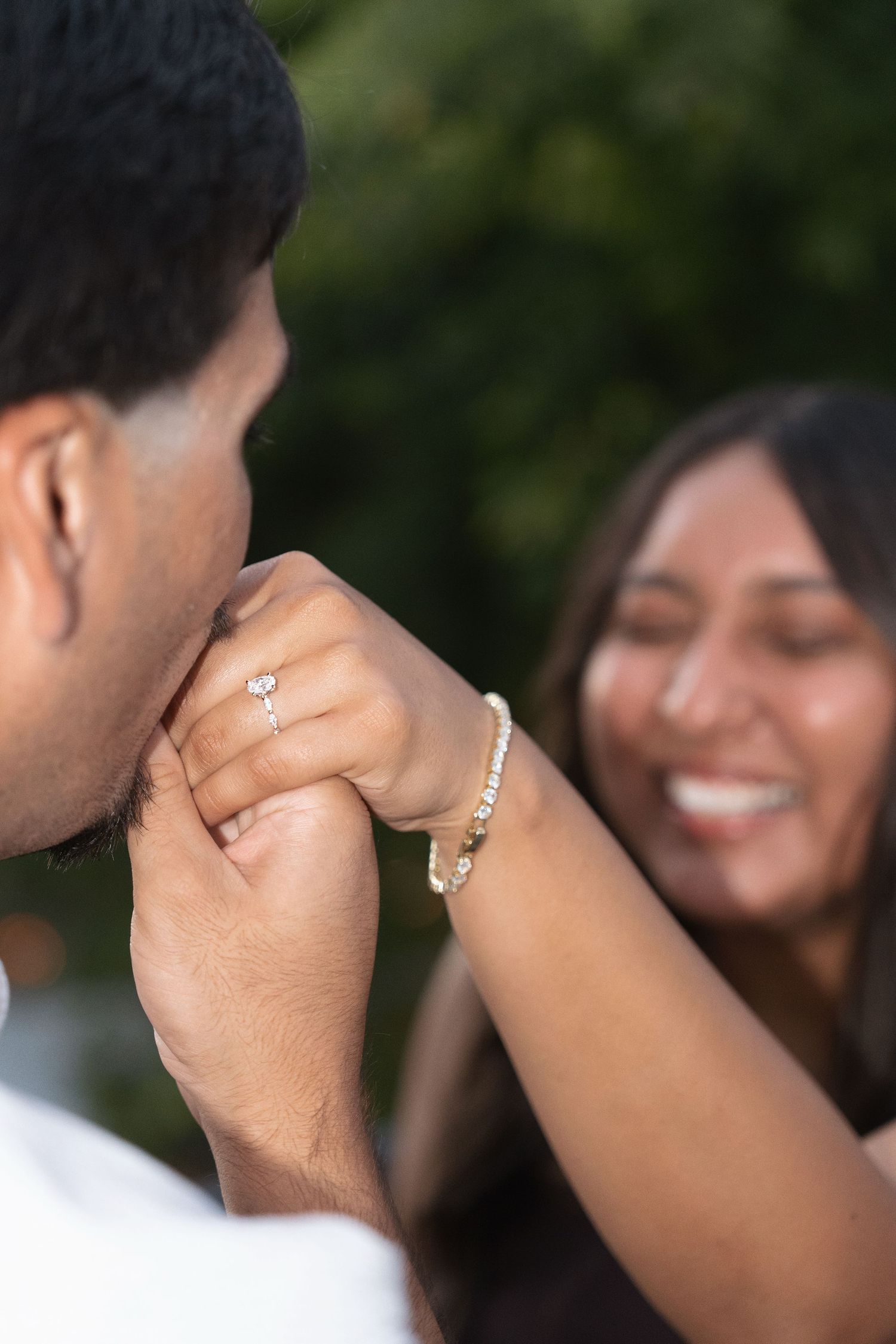 Man kissing woman's hand showing engagement ring while she smiles joyfully outdoors.