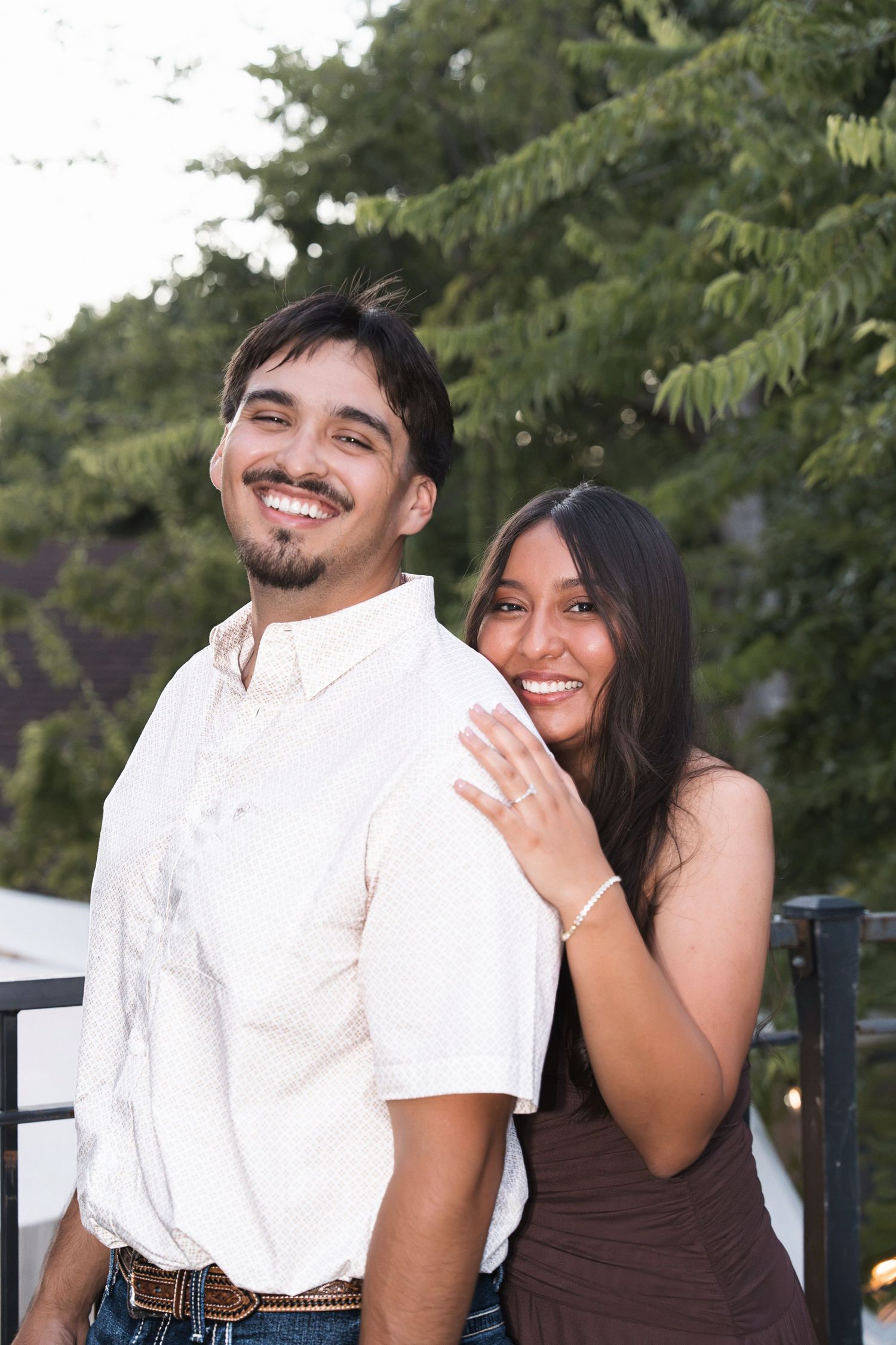 Smiling Hispanic couple posing outdoors, woman in brown dress showing engagement ring while embracing man in white shirt.