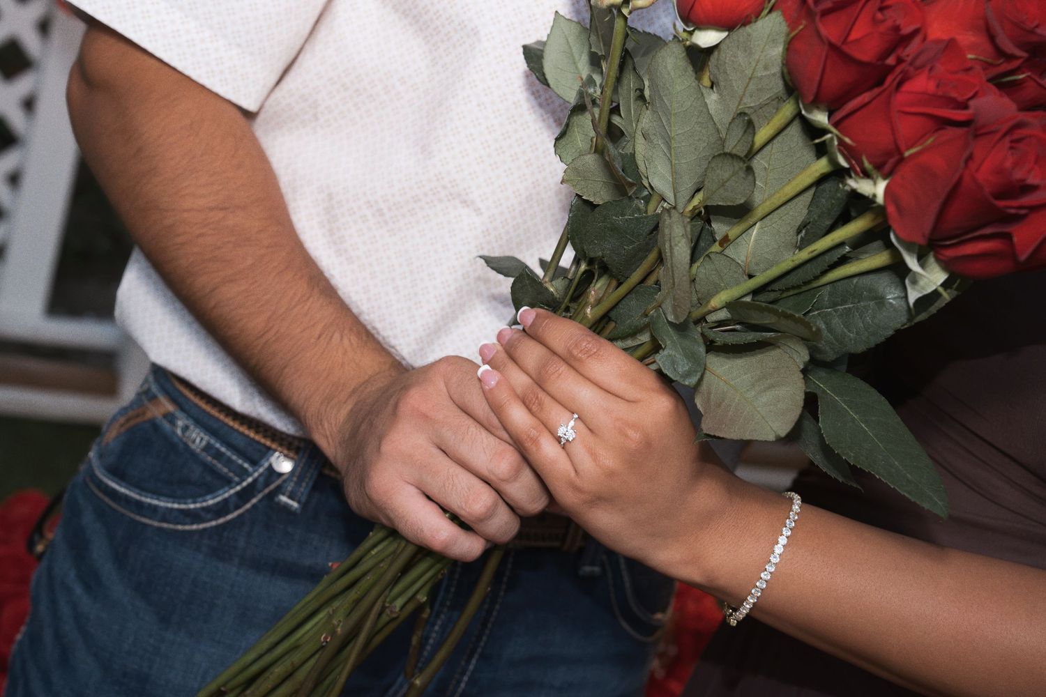 Close-up of engaged couple holding red roses bouquet with sparkling diamond engagement ring.