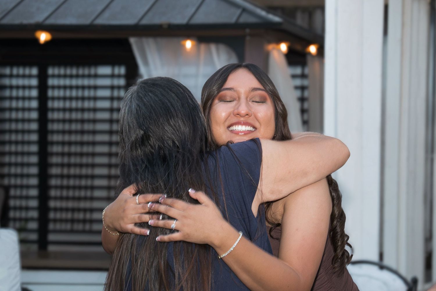 Two women sharing a warm embrace and smiling at an outdoor gathering, one wearing a brown dress with a bracelet.