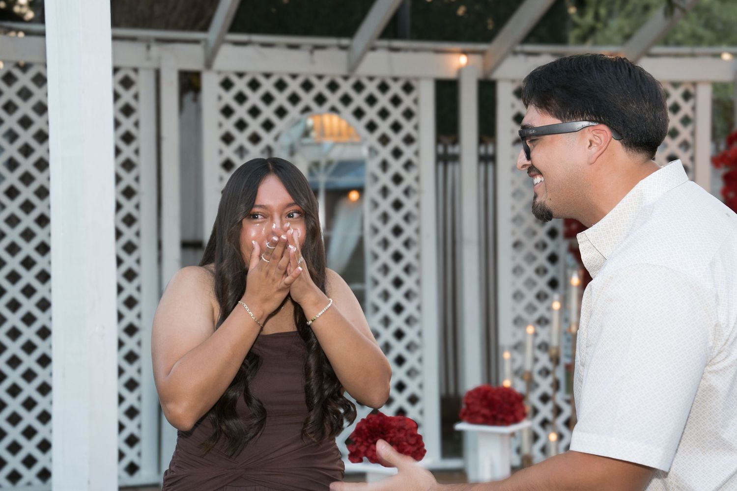 A man surprises a woman with a marriage proposal, holding red roses as she covers her mouth in shock outdoors.