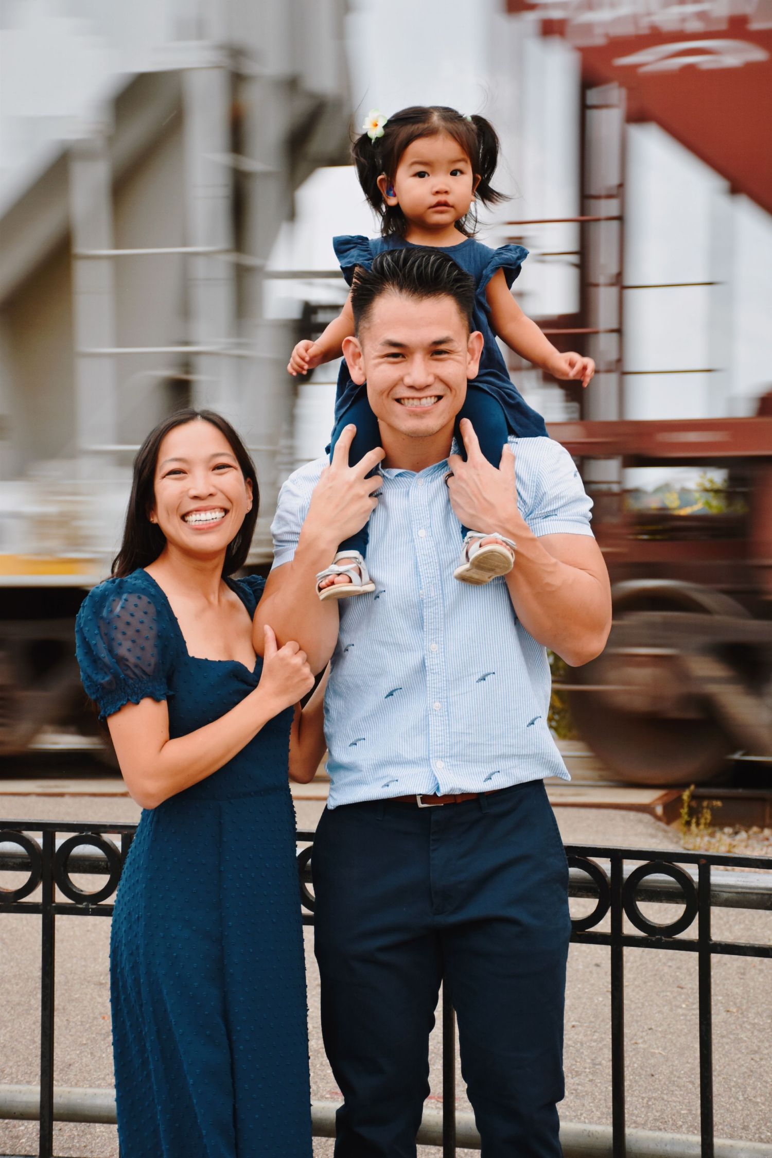 A smiling Asian family poses outdoors, with a young girl sitting on her father's shoulders while mother stands beside them.
