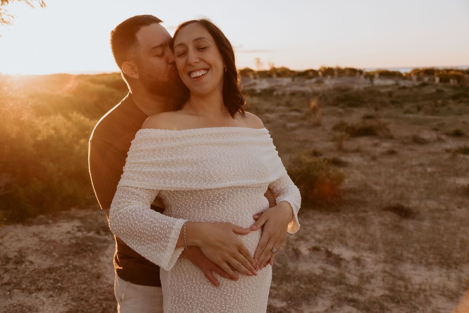 Couple sharing a tender maternity moment at golden hour sunset, man kissing woman's cheek while cradling her baby bump.