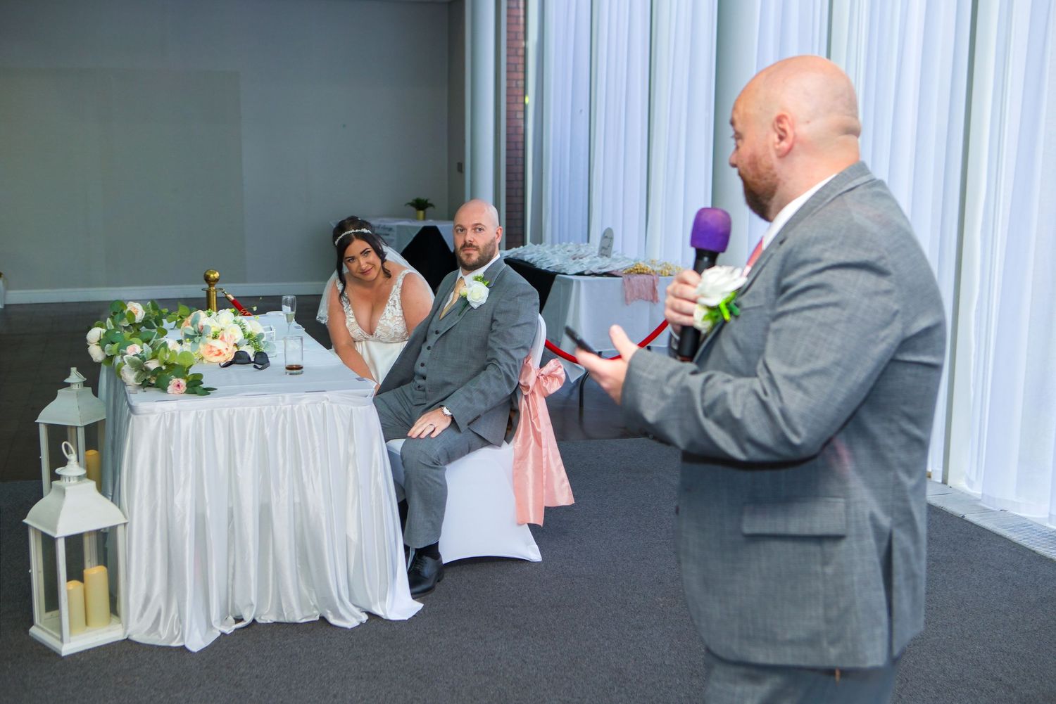 A bald man in a gray suit gives a toast speech to a bride and groom seated at a sweetheart table at a wedding reception.