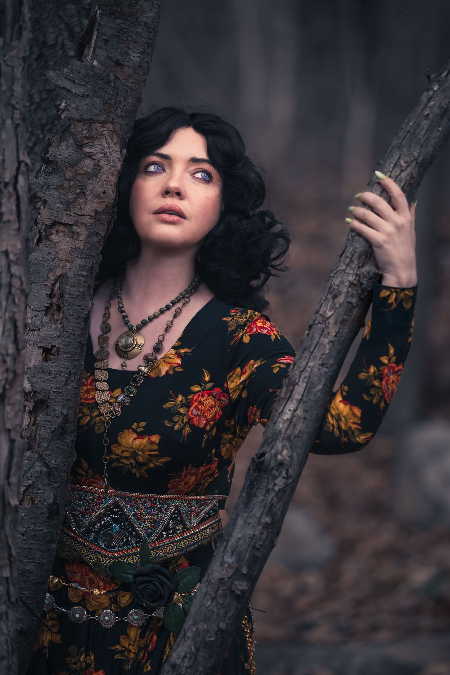Dark-haired woman in floral bohemian dress leaning against tree trunks in moody forest setting.