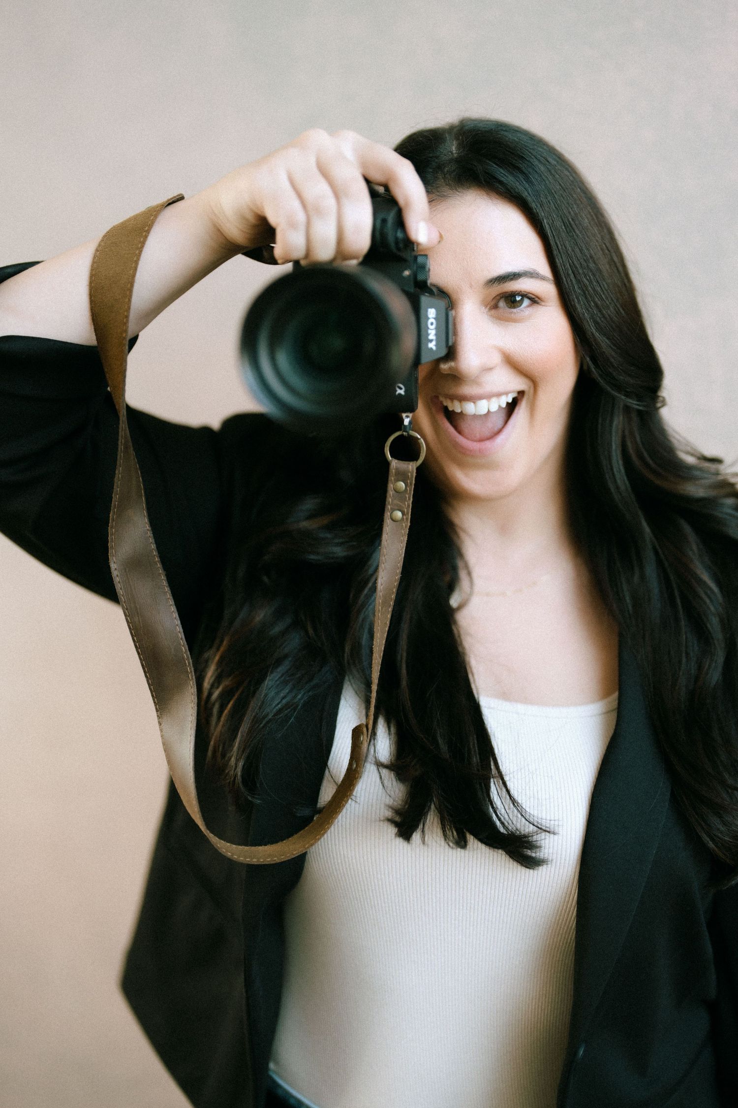 Excited brunette photographer smiling while holding Sony camera up against neutral beige background.
