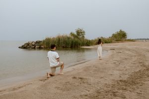A man kneels on one knee on a sandy beach while a woman in a white dress stands near the calm shoreline on a hazy, overcast day.