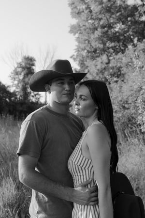 Black and white portrait of a couple embracing in a grassy field, the man in a cowboy hat and the woman in a striped sundress.