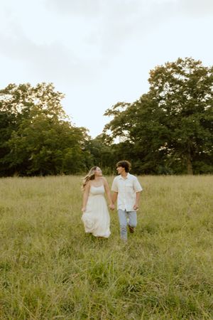 Couple holding hands and walking through a tall grassy meadow at sunset, with lush green trees in the background.