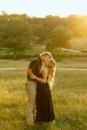 Couple embracing and kissing in a grassy field at golden hour sunset, with trees and a winding path in the background.