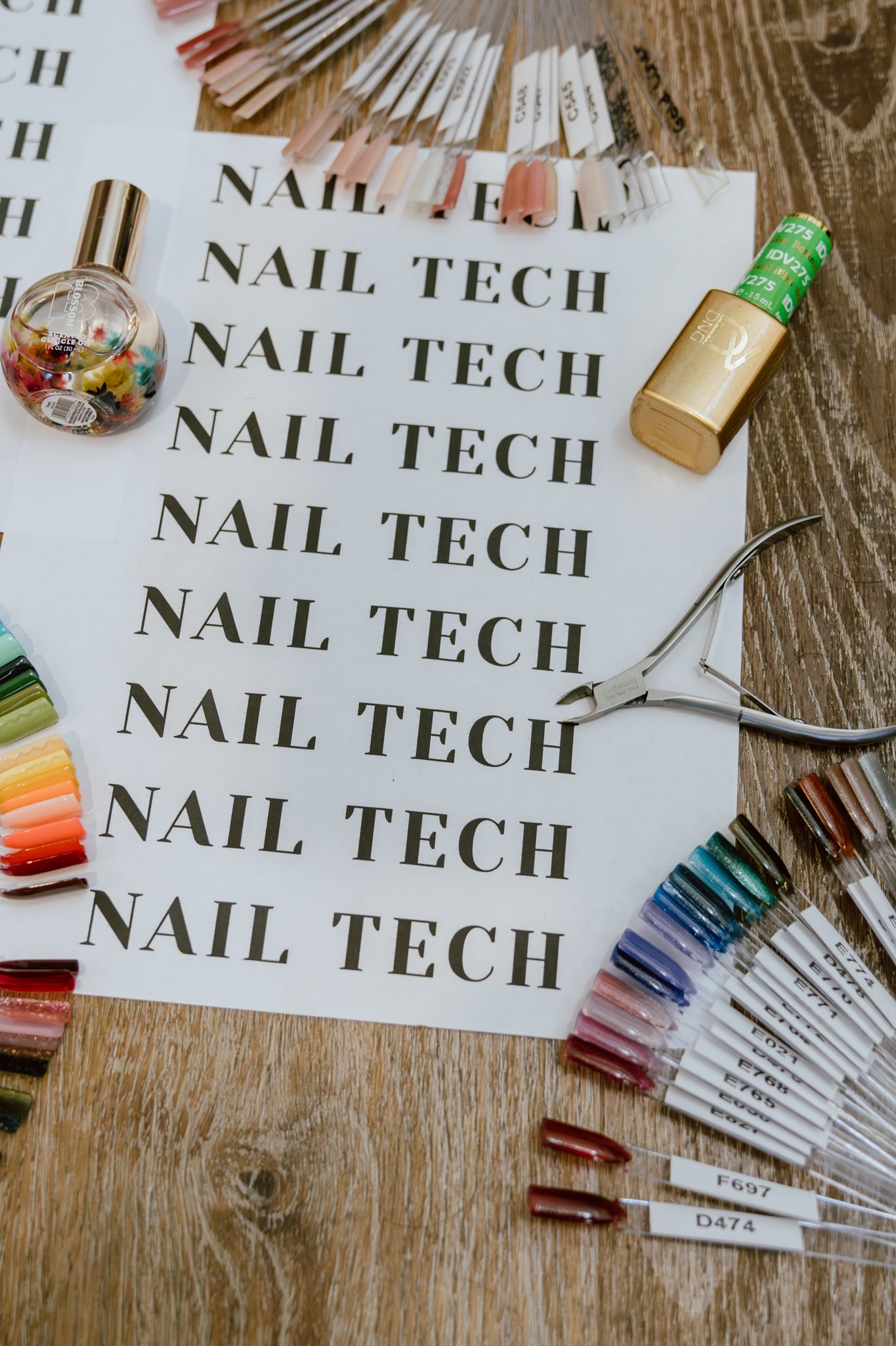 Nail tech workspace with cuticle nippers, gel polish swatch sticks, cuticle oil and a polish bottle on a paper printed with NAIL TECH.