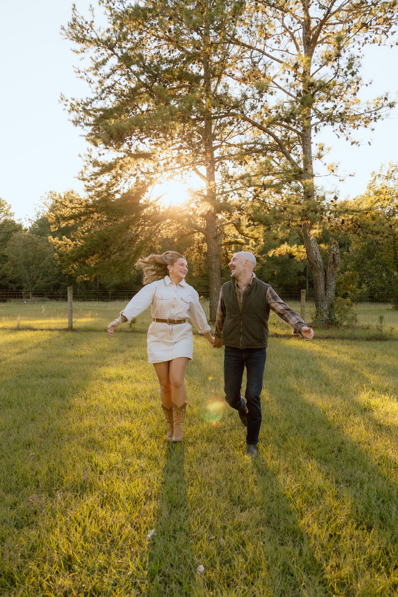 Happy couple holding hands and running through a sunlit grassy field with tall trees and golden sunset light behind them.