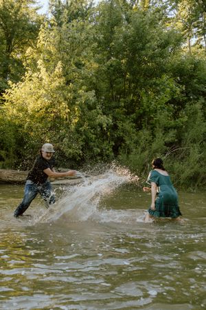 Man in a cap and woman in a green dress splash water at each other while playing in a shallow river surrounded by lush green trees.