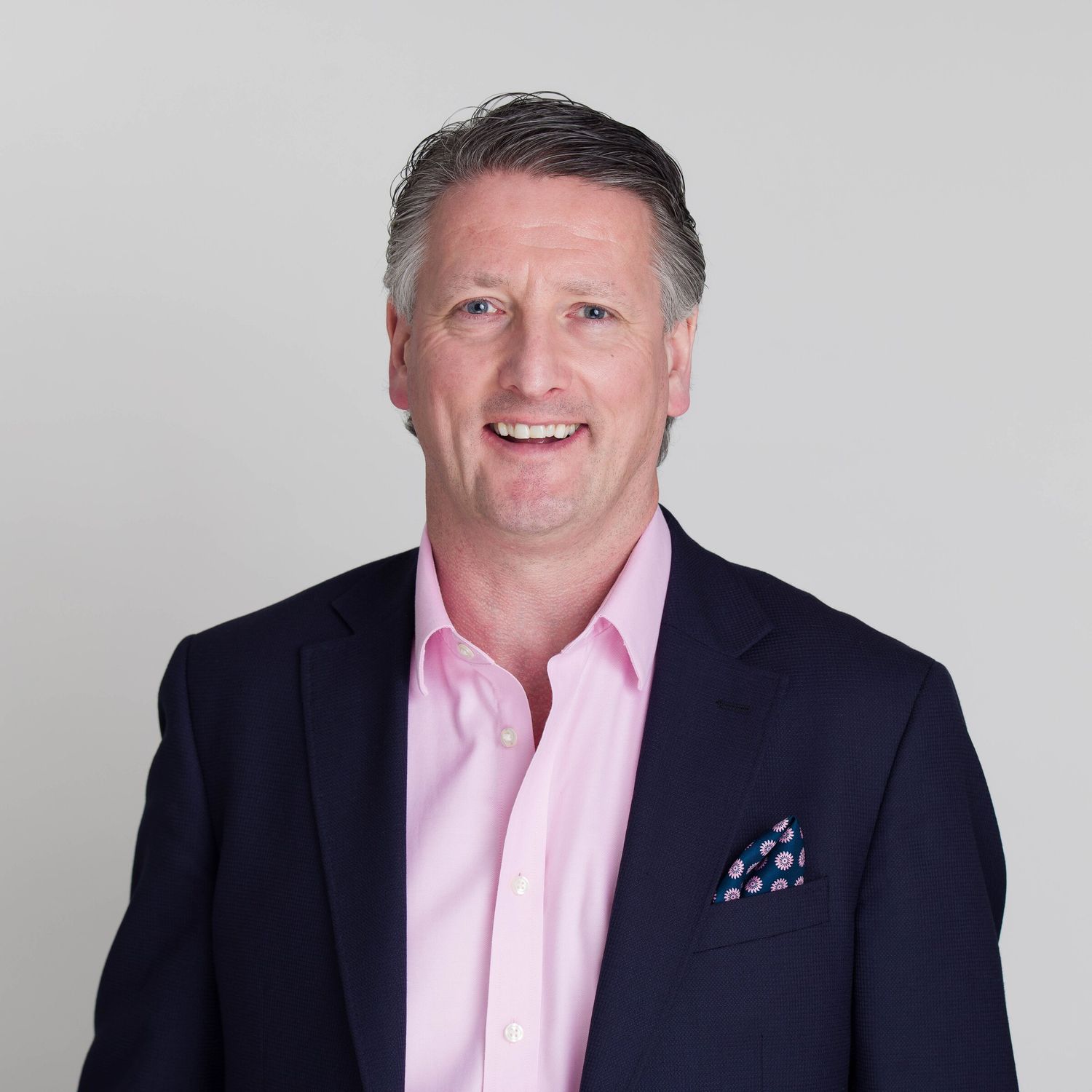 Relaxed corporate portrait of a businessman in a navy blazer and pink shirt on a pale background
