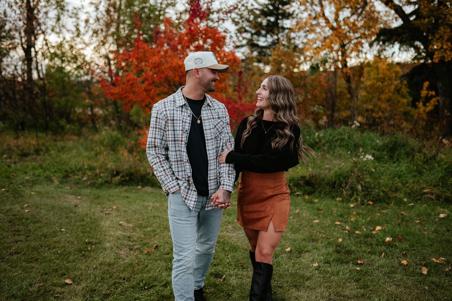 Smiling couple holding hands and walking together on a grassy path surrounded by colorful autumn trees during a fall engagement photoshoot.