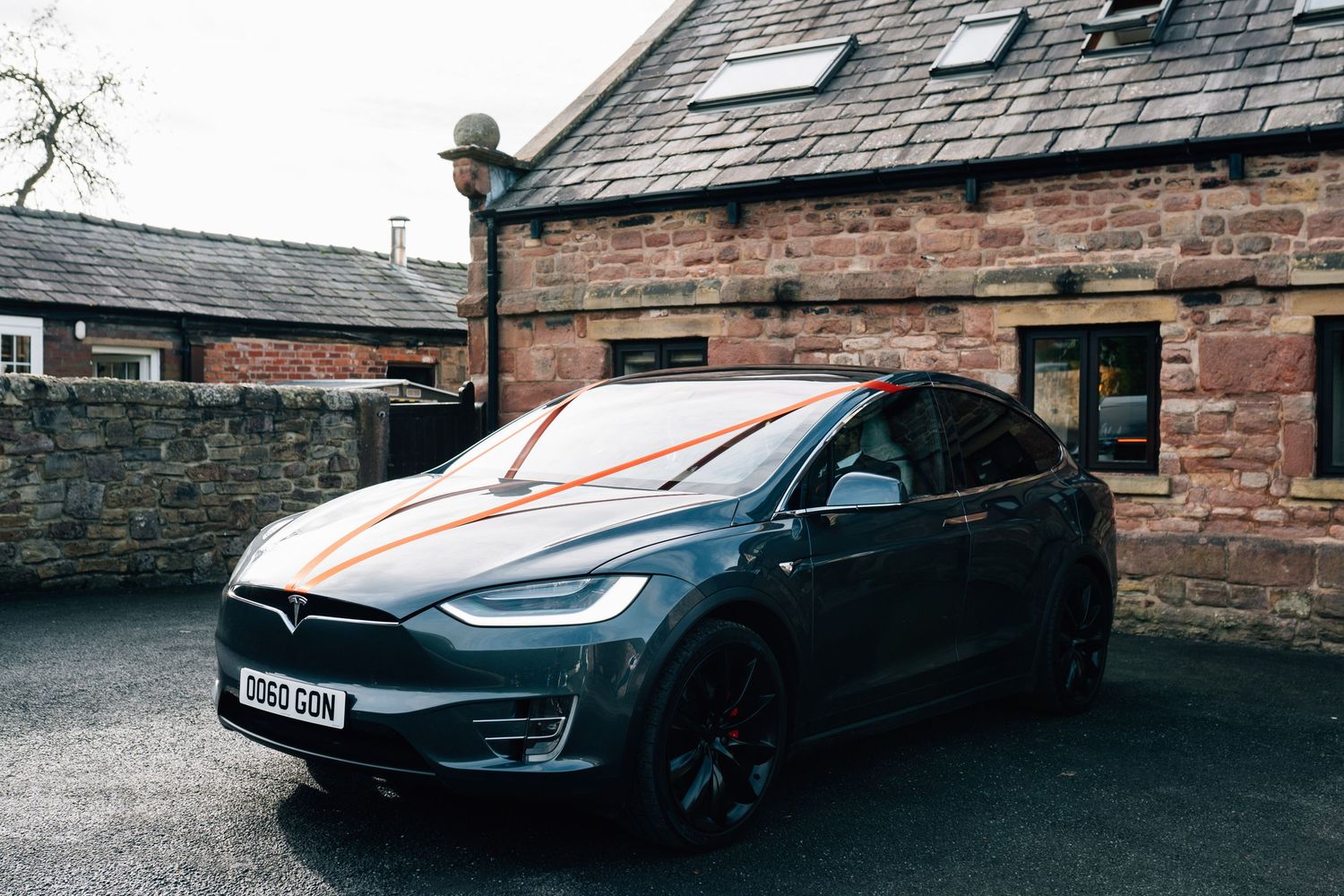Dark gray Tesla Model X electric SUV with orange reflections parked on a driveway beside a rustic red sandstone building with a slate roof.