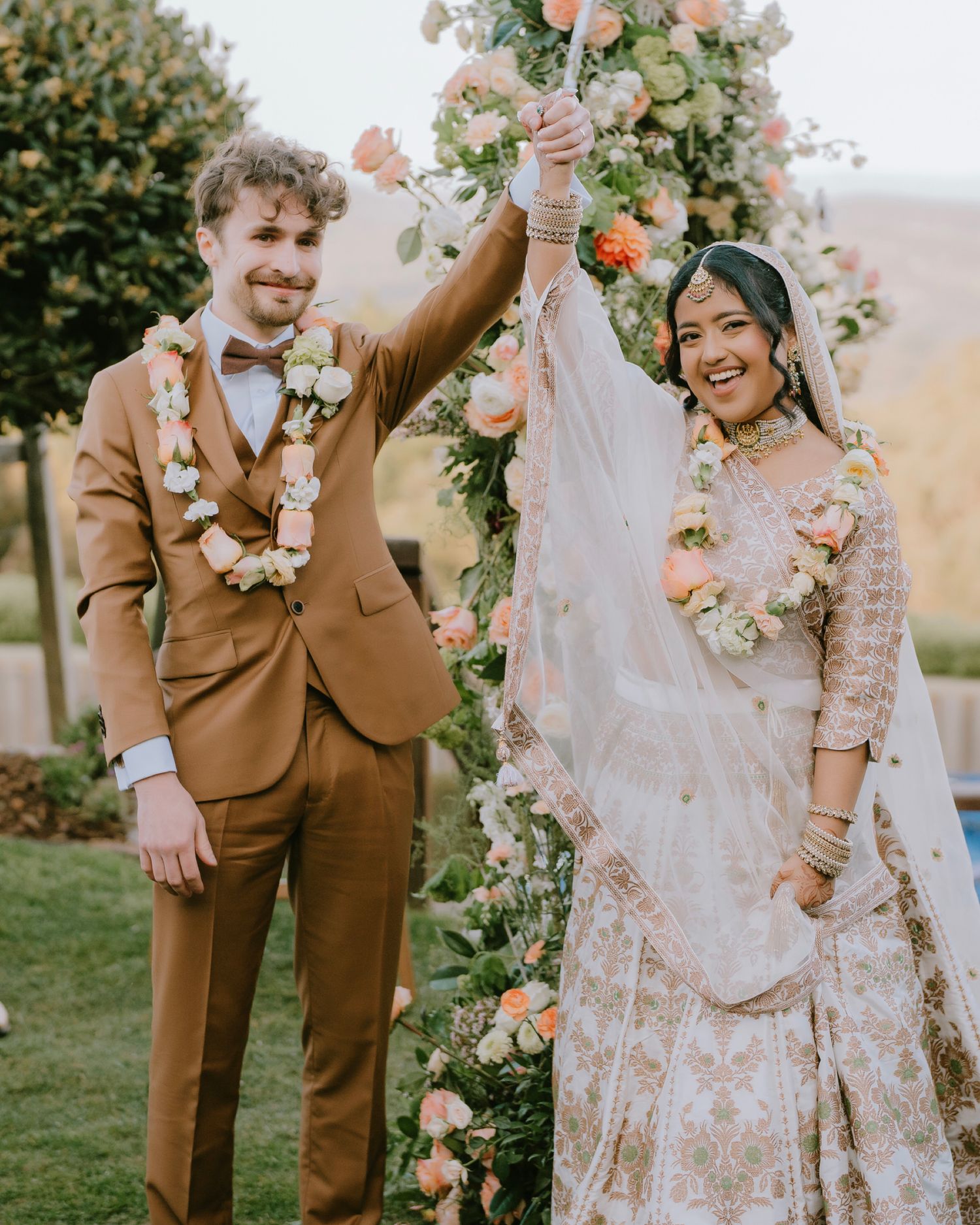Couple in floral garlands raise joined hands under a rose arch - Adelaide South Asian wedding ceremony photos