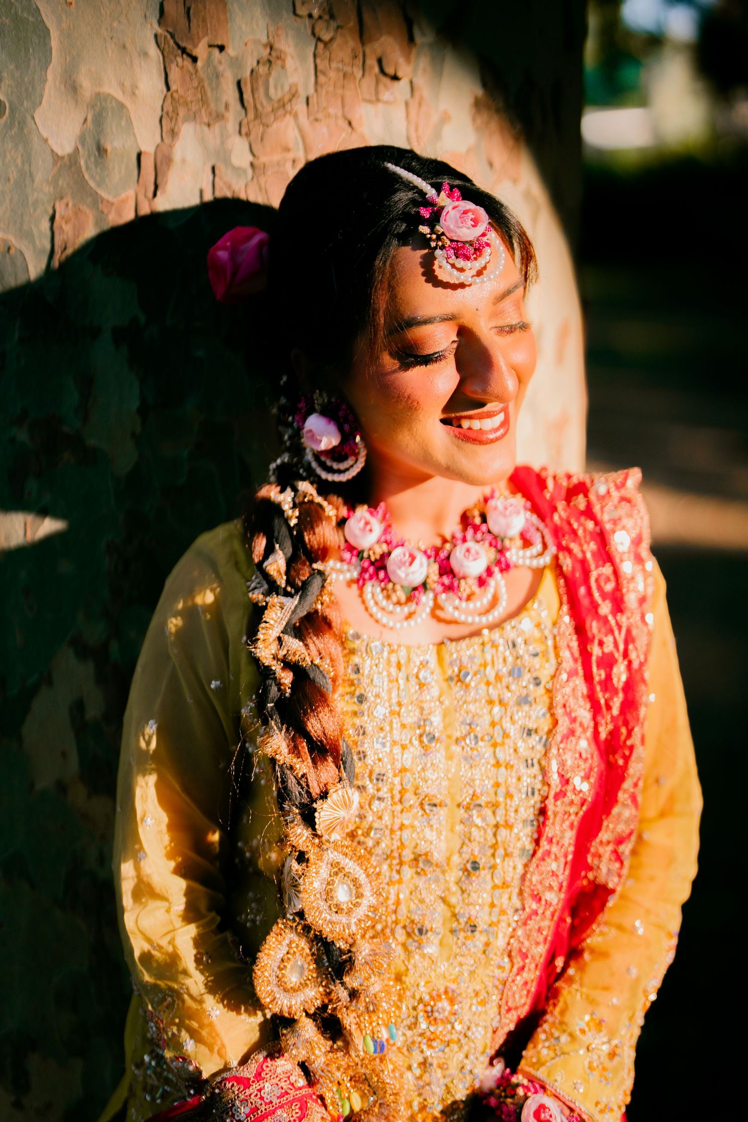 Smiling bride in a yellow and red embroidered outfit with floral jewelry and a long braid, leaning against a tree in warm sunlight.