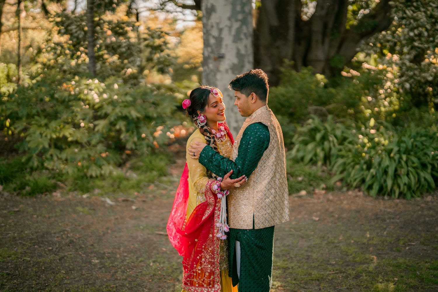 Couple in traditional Indian wedding attire embrace in a sunlit garden, the bride in a red and gold sari with floral hair accessories.