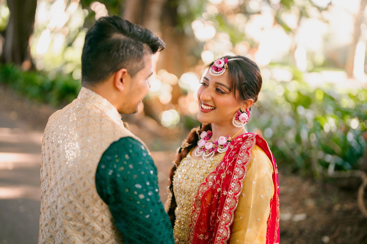 Smiling woman in gold and red traditional Indian attire with floral hair jewelry laughs with a man in a cream vest and green kurta outdoors.