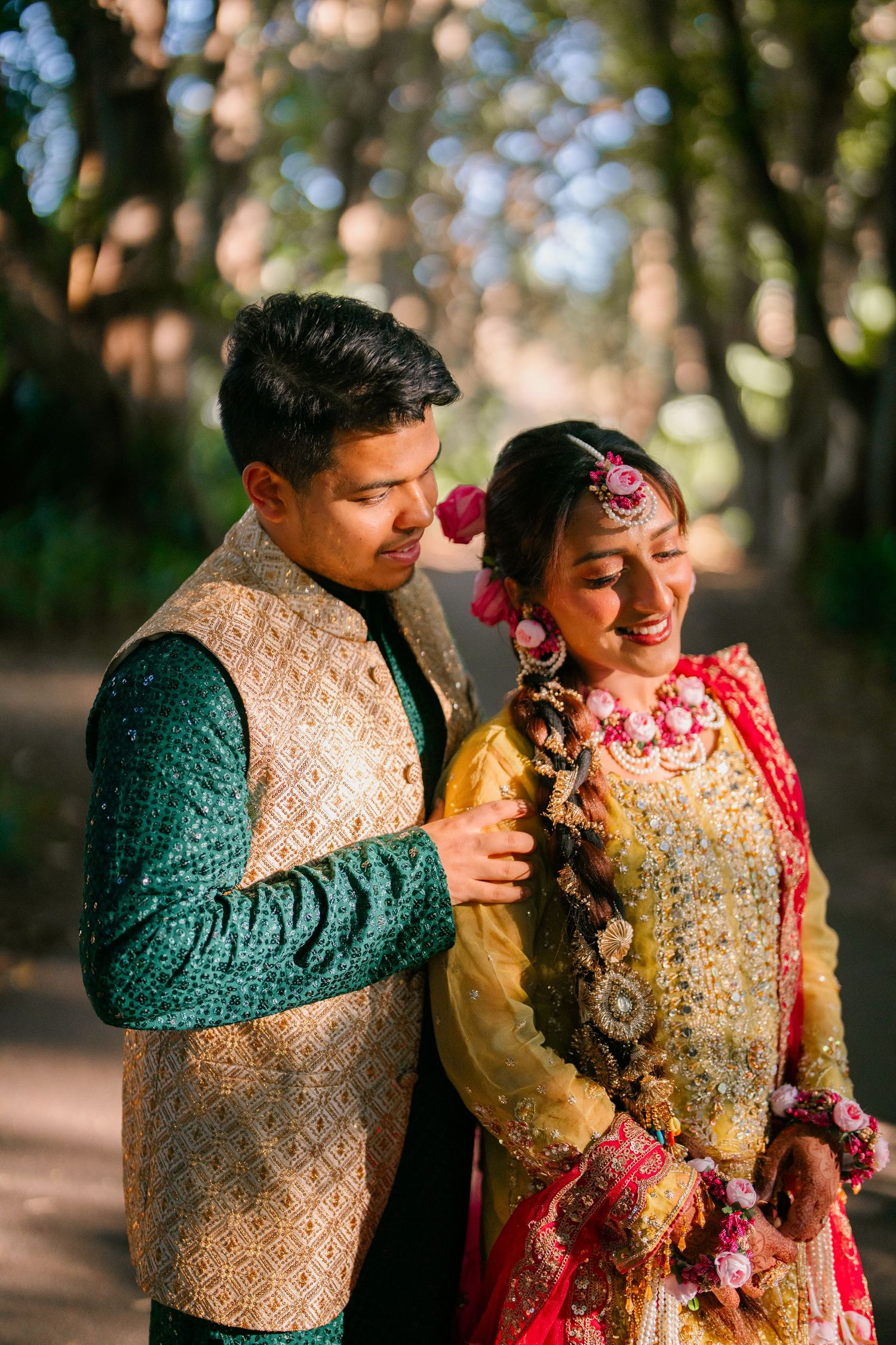 Smiling couple in traditional South Asian wedding attire, he in a green and gold vest, she in a gold and pink outfit, posing outdoors.