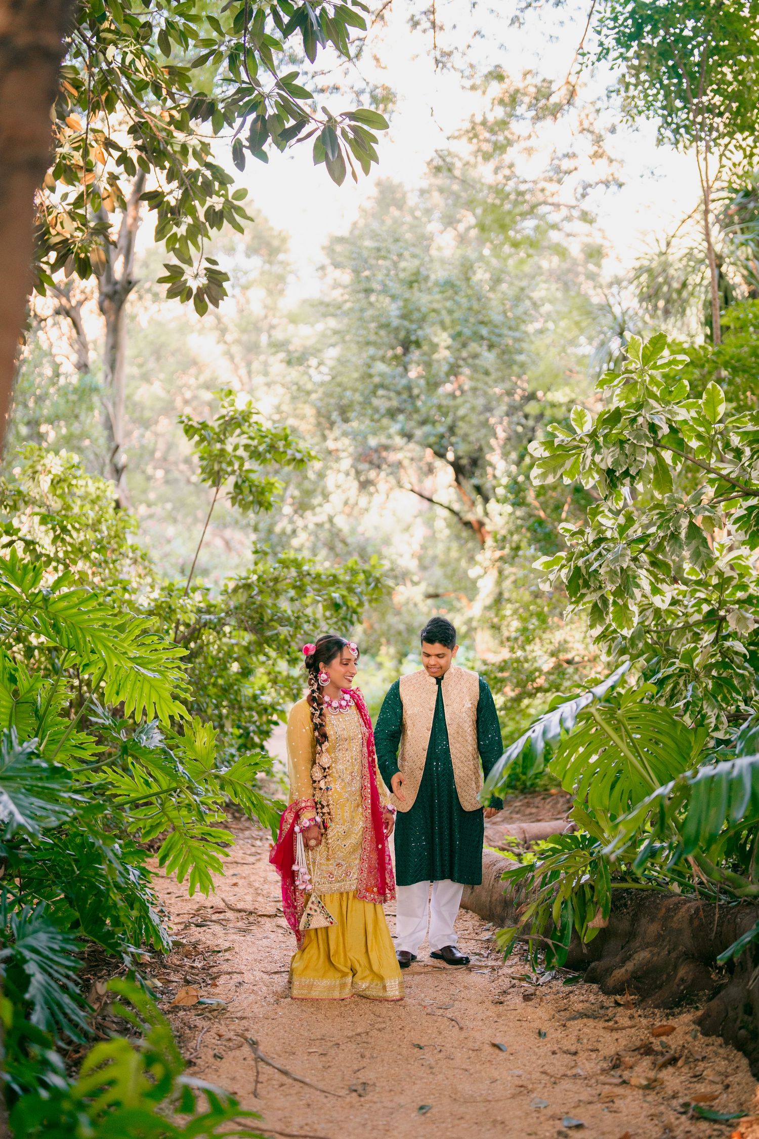 A couple in traditional Indian wedding attire, she in yellow with a red dupatta, he in green, walk a dirt path through lush green trees.