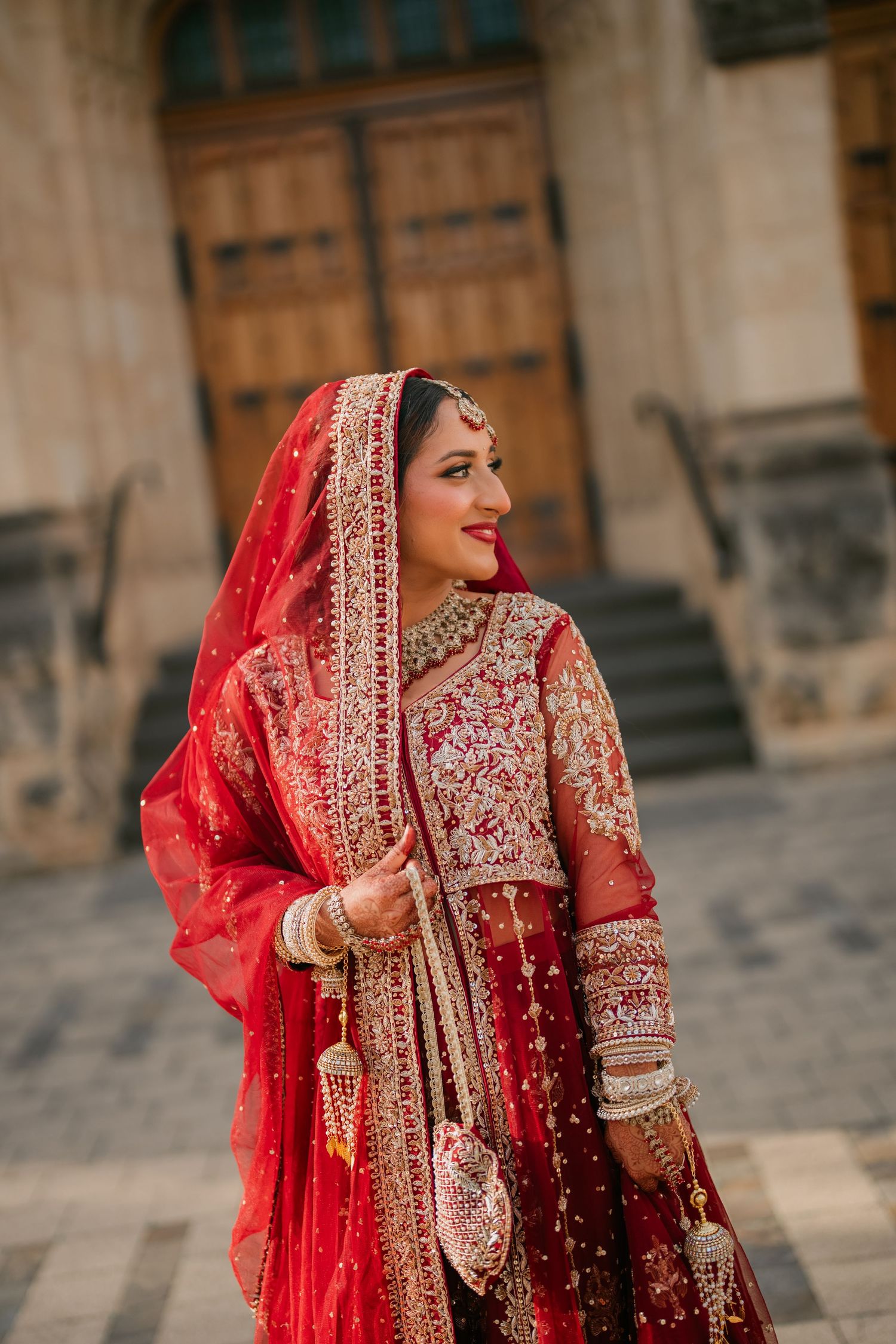 Smiling bride in a red embroidered lehenga with dupatta, gold jewelry and bangles posing outside a historic building with wooden doors.