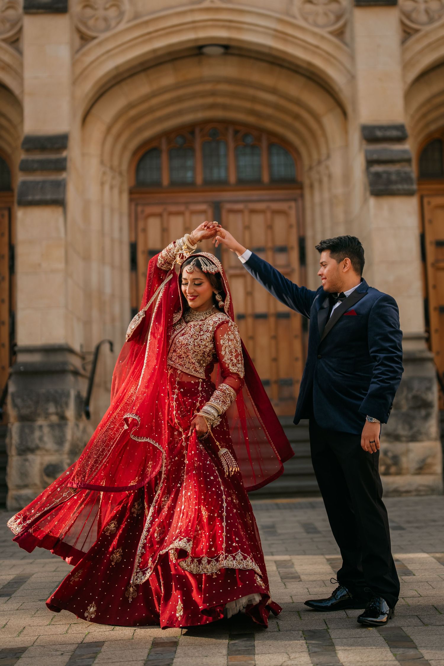 Bride in a red embroidered lehenga and dupatta twirls with her groom in a navy suit outside a grand stone arched building.