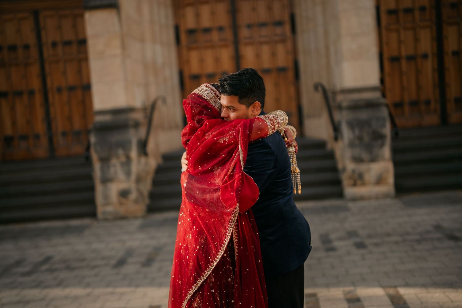 Bride in a red embroidered dupatta embraces a groom in a dark suit in front of a historic building with wooden doors.