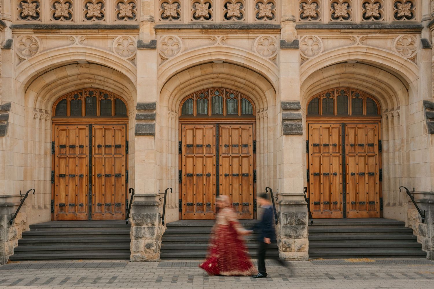 Bride in a flowing red gown walks beside a groom in a dark suit past grand arched wooden doors and stone steps of a historic building.