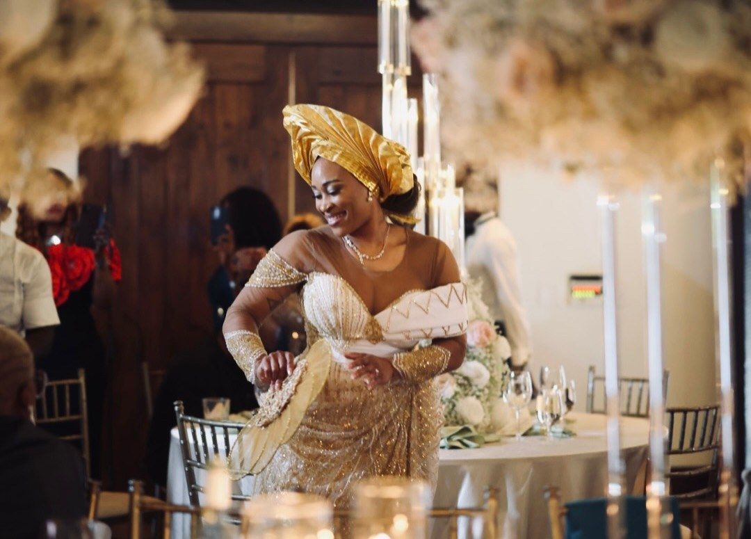 Smiling woman in a gold gele headwrap and embellished gold gown stands among elegant candlelit tables at a wedding reception.