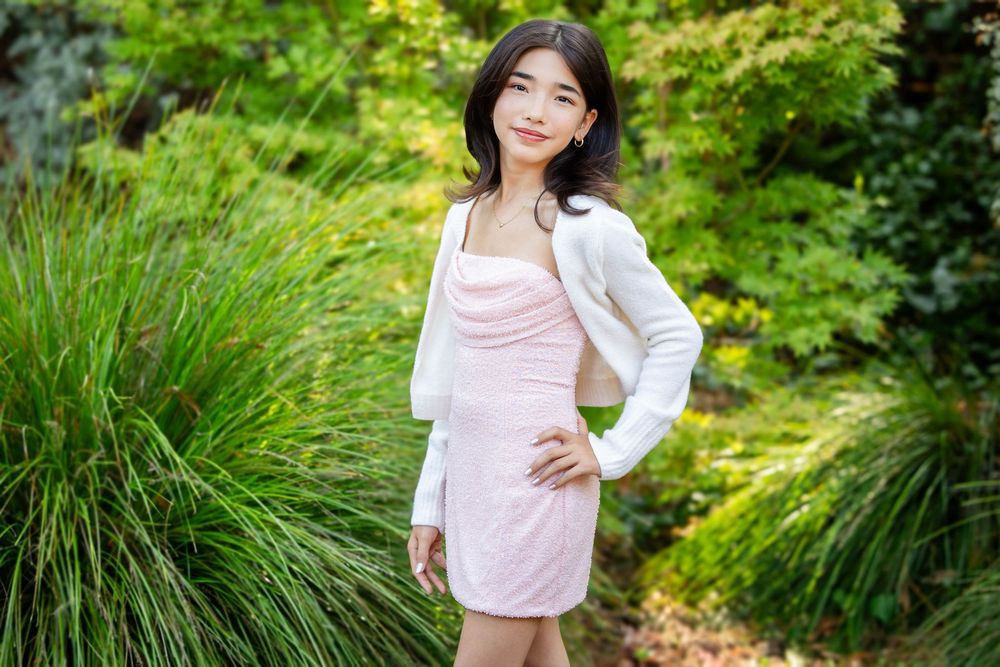 Young girl in a pink satin dress and white cardigan poses with hands on hips among lush green plants in a sunny garden.