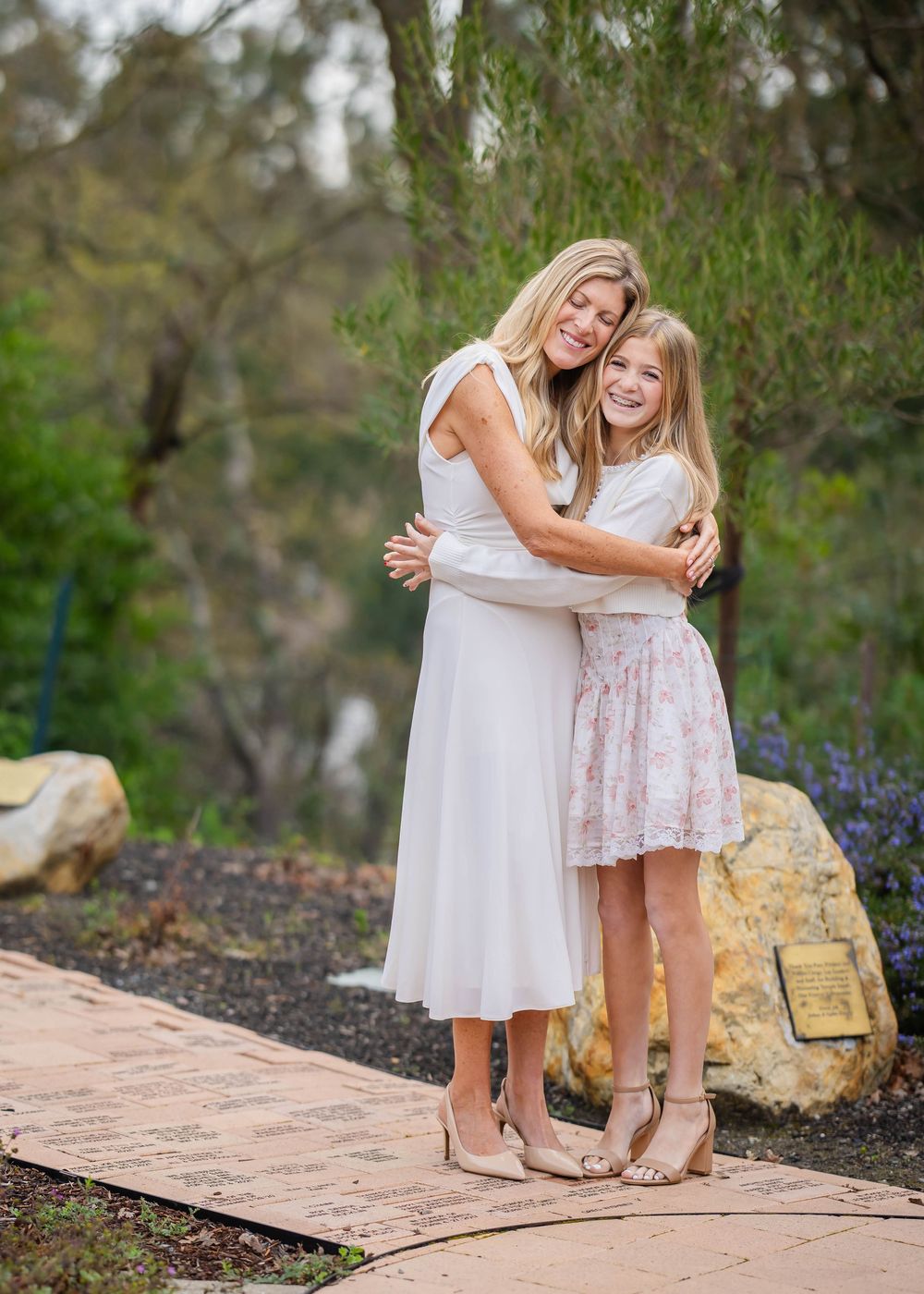 Two smiling women in light dresses and heels embrace on a brick pathway in a leafy garden beside large boulders and purple flowers.