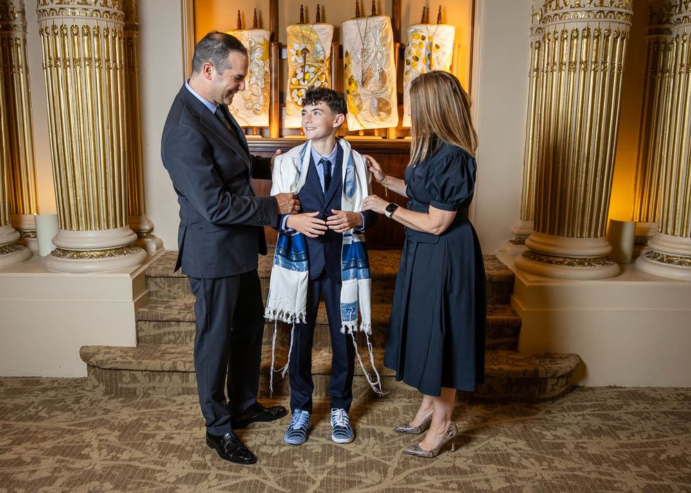 Parents stand beside a smiling boy wearing a prayer shawl during a bar mitzvah celebration in an elegant hall with ornate gold columns.