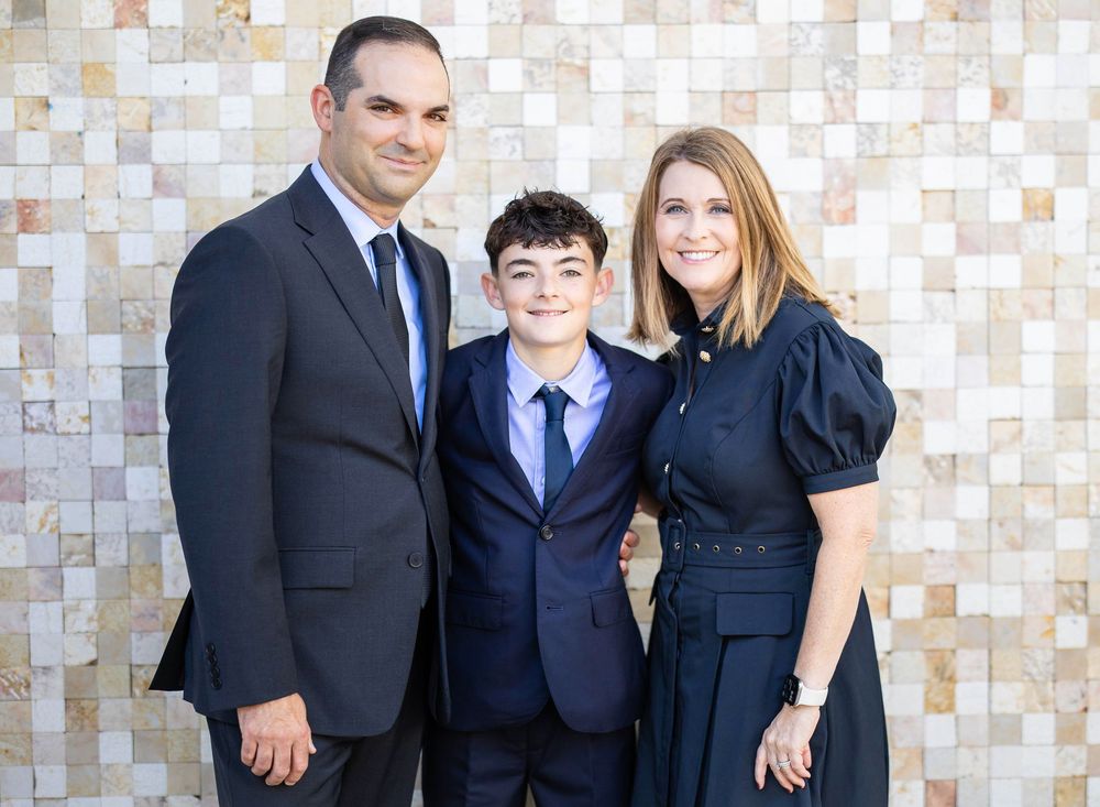 Smiling family of three in formal attire, a man in a dark suit, a boy in a navy suit and a woman in a navy dress, posing against a tiled wal