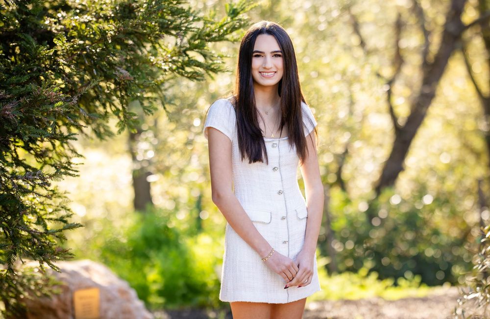 Smiling young woman with long dark hair in a white button-front dress standing outdoors among sunlit trees and greenery.